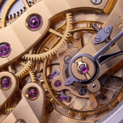 Das Uhrenlexikon zeigt anhand einer Nahaufnahme komplizierter Uhrzahnräder und -mechanismen verschiedene goldfarbene Zahnräder, Federn und Schrauben. In einigen Teilen sind winzige rosa Edelsteine eingelassen, die das komplexe Innenleben einer mechanischen Uhr veranschaulichen.