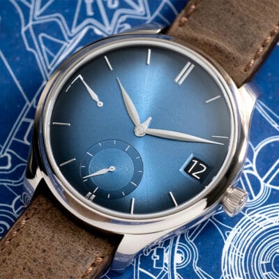 Eine Luxusarmbanduhr von Uhrenlexikon mit poliertem Edelstahlgehäuse, blauem Sunburst-Zifferblatt und braunem Lederarmband. Das Zifferblatt verfügt über silberne Zeiger, ein Datumsfenster auf der 3-Uhr-Position und Hilfszifferblätter. Die Uhr ist elegant auf einer blau gemusterten Oberfläche platziert.