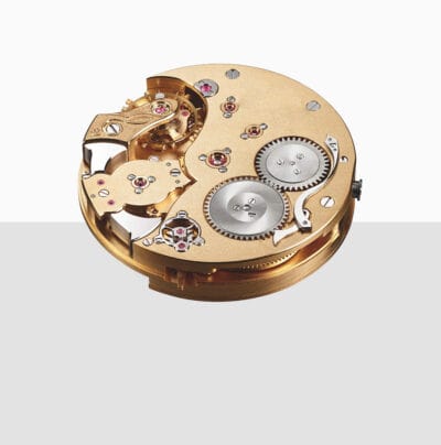 Eine Nahaufnahme eines mechanischen Uhrwerks mit komplizierten goldenen Zahnrädern, Rubinen und silbernen Komponenten, die vor einem weiß-grauen Hintergrund mit Farbverlauf sichtbar sind.