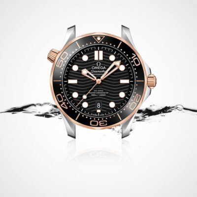 Eine Luxusarmbanduhr mit schwarzem Zifferblatt, goldenen Akzenten und einer schwarz-goldenen Lünette mit dem Markennamen „Omega Seamaster“. Die Uhr ist vor einem weißen Hintergrund mit Spritzwasser abgebildet, was ihre wasserdichte Eigenschaft unterstreicht. Weitere Details finden Sie im Uhrenlexikon.