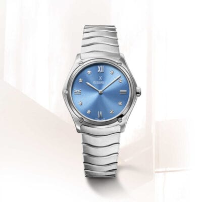 Eine elegante, silberne Armbanduhr mit einem metallisch blauen Zifferblatt. Die Uhrenlexikon-Uhr verfügt über römische Ziffern an den Positionen 12, 3, 6 und 9 sowie diamantartige Markierungen für die restlichen Stunden. Das Band hat ein elegantes, wellenförmiges Design. Der Hintergrund ist sanft beleuchtet und minimalistisch.