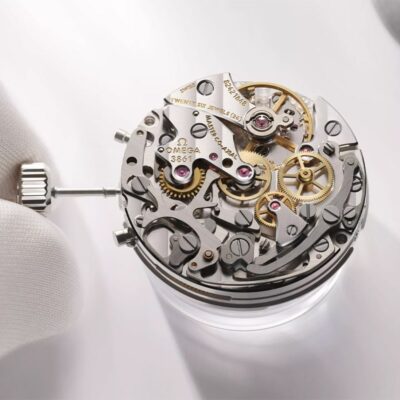 Nahaufnahme eines detaillierten Uhrwerks, das von einer Person mit weißen Handschuhen bedient wird. Die komplizierten Zahnräder, Zahnräder und Komponenten, einige mit der Aufschrift „OMEGA“ und „26 JEWELS“, sind sichtbar, während sie vom Experten anhand seines Uhrenlexikons sorgfältig freigelegt und eingestellt werden.