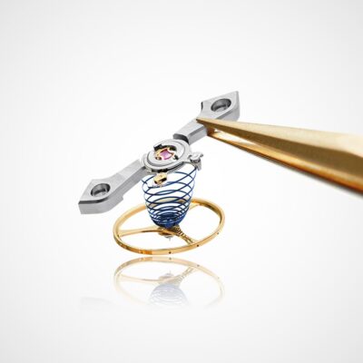 Nahaufnahme der Unruh und des Hemmungsmechanismus eines Uhrwerks, das im Uhrenlexikon erwähnt wird und mit einer Pinzette gehalten wird. Die Baugruppe besteht aus einer gewickelten blauen Spiralfeder, einem goldfarbenen Ring und einem Metallarm mit zwei Löchern. Die Komponenten sind vor einem schlichten weißen Hintergrund sehr detailliert dargestellt.